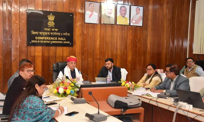 Bhagirath Choudhary, in a red turban, chairs a meeting at a long table in the wood-panelled conference hall of the Office of the District Commissioner, Nagaon, with officials on either side