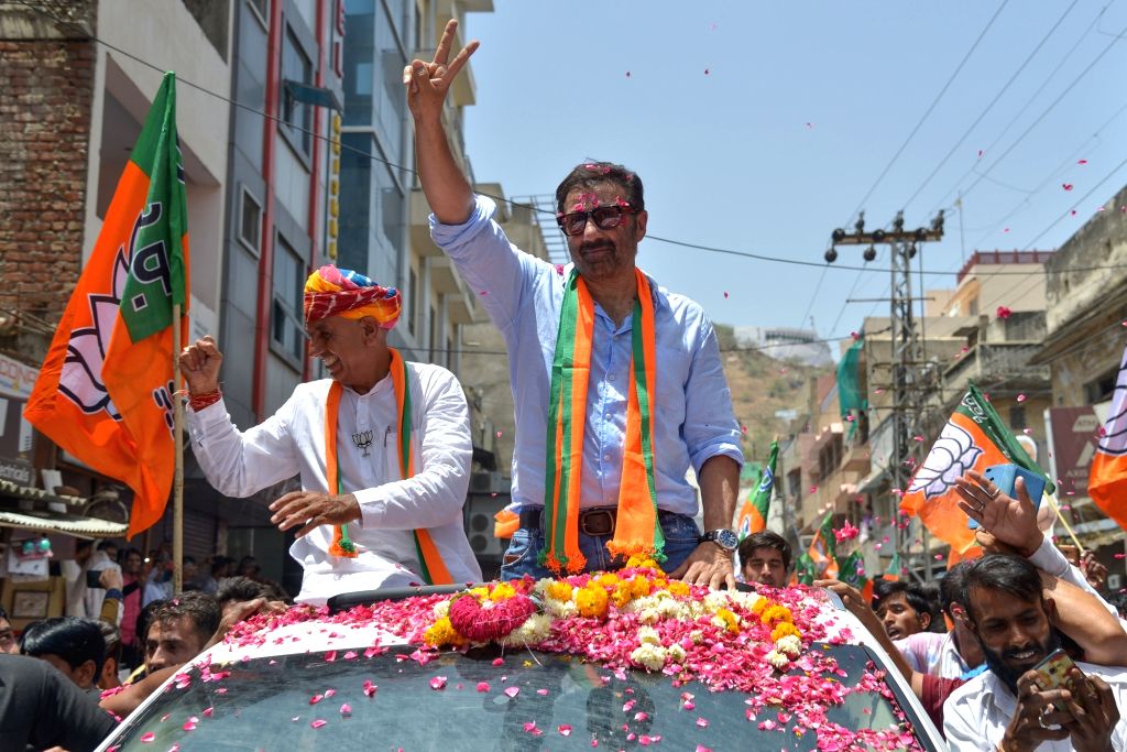 Bhagirath Choudhary and Sunny Deol wave from a flower-covered open vehicle during a roadshow